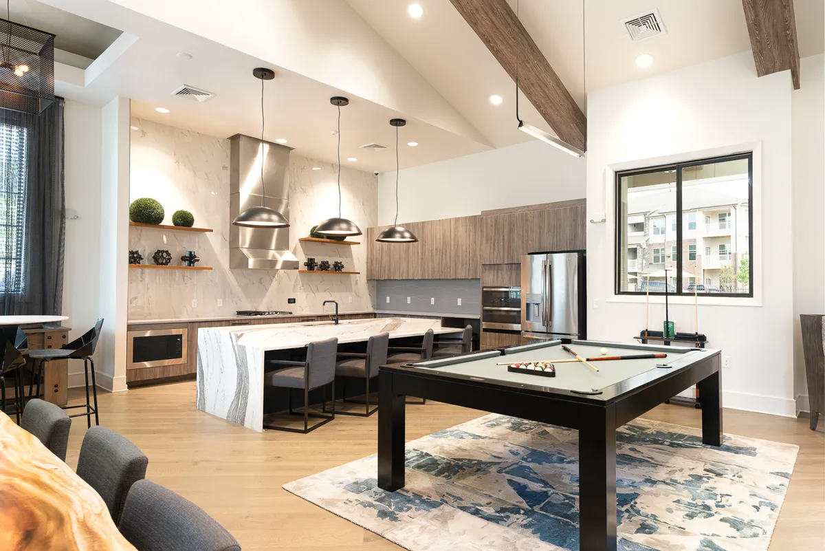 An open-plan space balancing a kitchen island, dining area, and pool table, illuminated by large windows. The kitchen island is central, ideal for meal prep or casual gatherings, while the proximity to the dining area and recreational space encourages communal interaction.