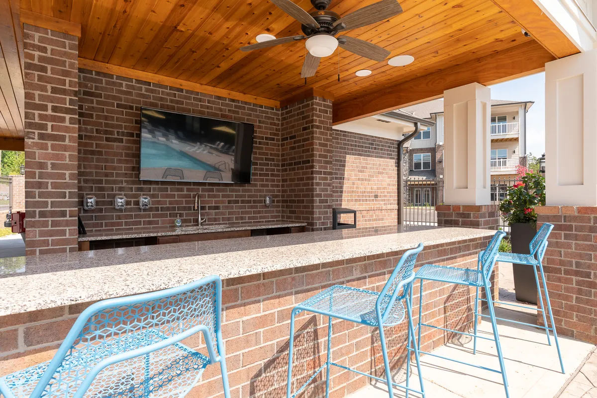 This cheerful exterior shot highlights a charming bar area with bright blue chairs, a mounted TV screen, and a brick wall backdrop. The space is designed for outdoor socializing, dining, and relaxing, suggesting opportunities for family gatherings and shared moments.