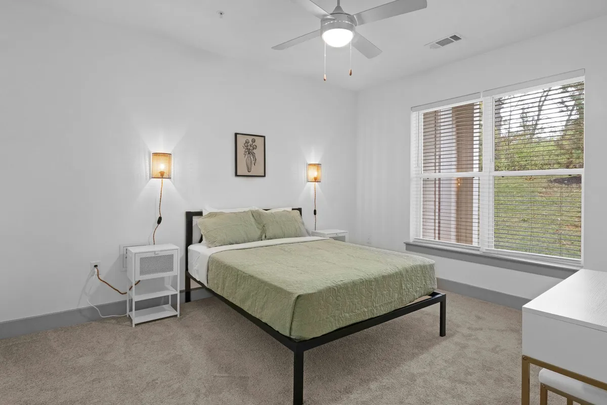 This serene bedroom features a large, inviting bed flanked by matching nightstands and topped with tasteful artwork. Sunlight streams through the windows, creating a tranquil, peaceful atmosphere perfect for unwinding.