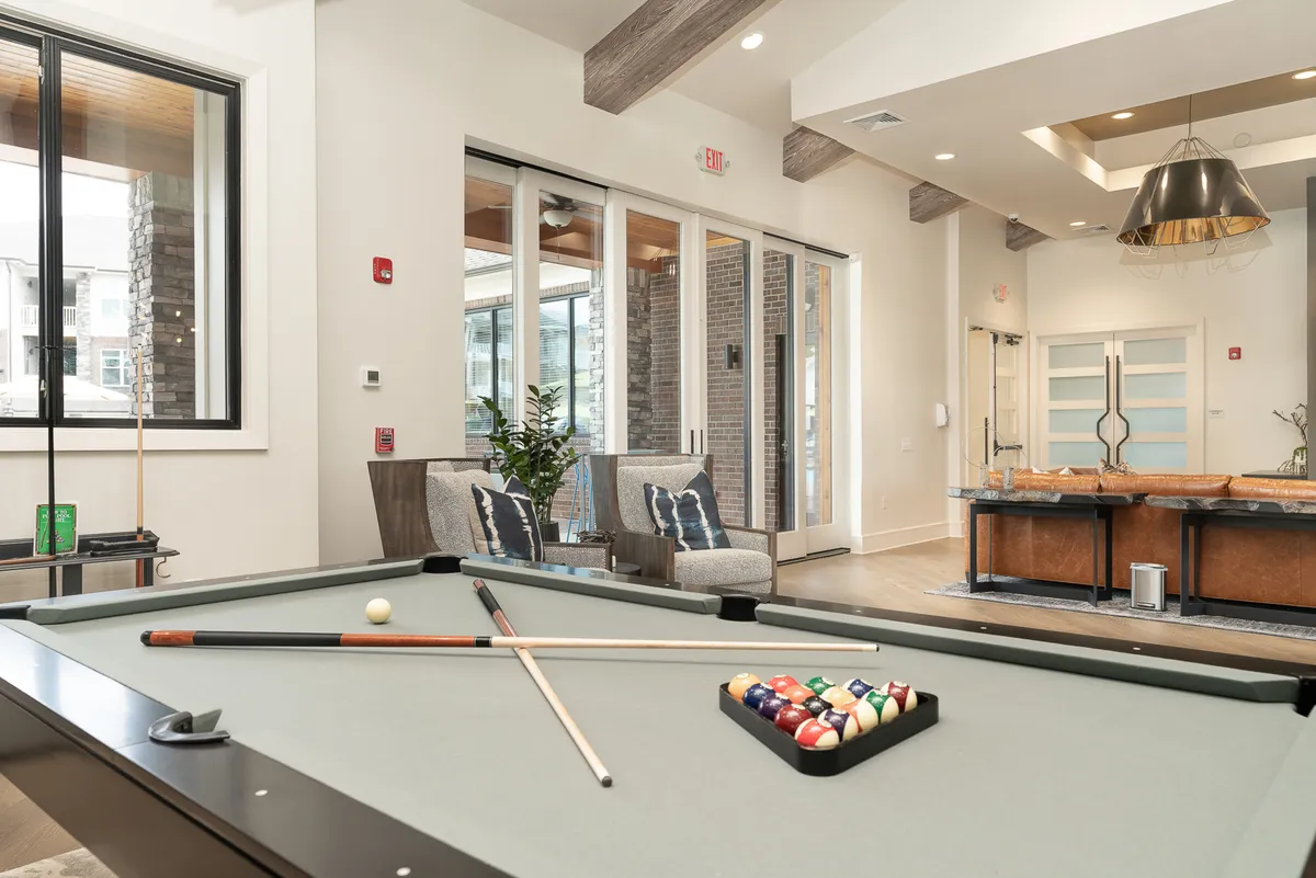 A stylish indoor lounge featuring a pool table and a comfortable seating area flooded with natural light from large windows. The arrangement encourages casual social interaction, whether playing billiards or relaxing with friends after exploring the city.