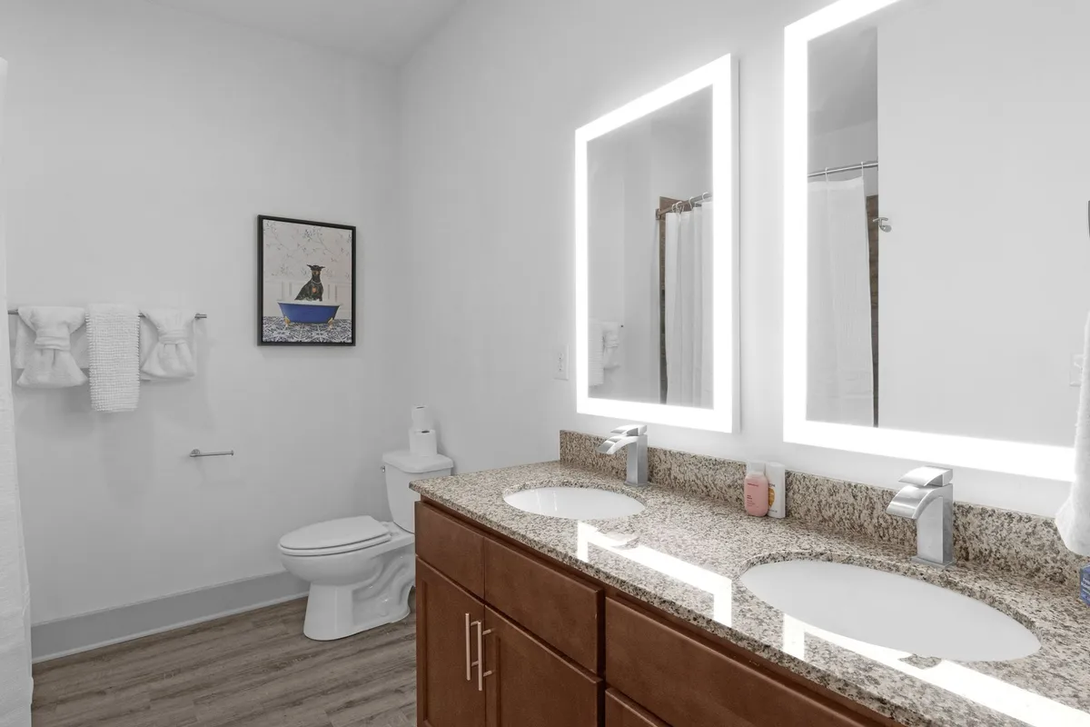 A spacious, modern bathroom featuring a double sink with sleek faucets, illuminated mirrors, and tasteful wall art. The fresh, inviting vibe offers glimpses of stylish fixtures and an atmosphere ideal for self-care routines.