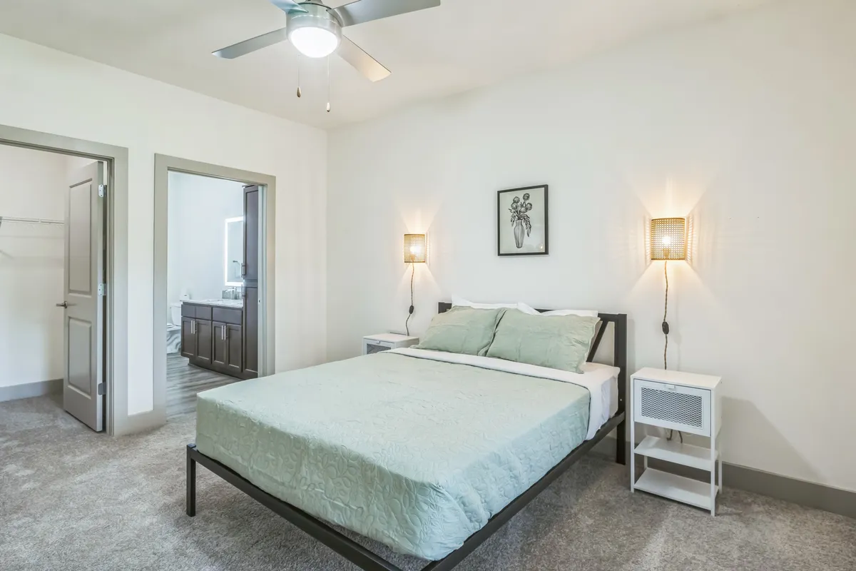 This image showcases a comfortable bedroom with a well-made bed, inviting wall art, and nightstands. The scene is softly lit with natural light filtering in, and a bathroom doorway is visible, hinting at the convenience of en-suite amenities. The overall mood is calm and cozy, reflecting a relaxing atmosphere ideal for long-term stays.