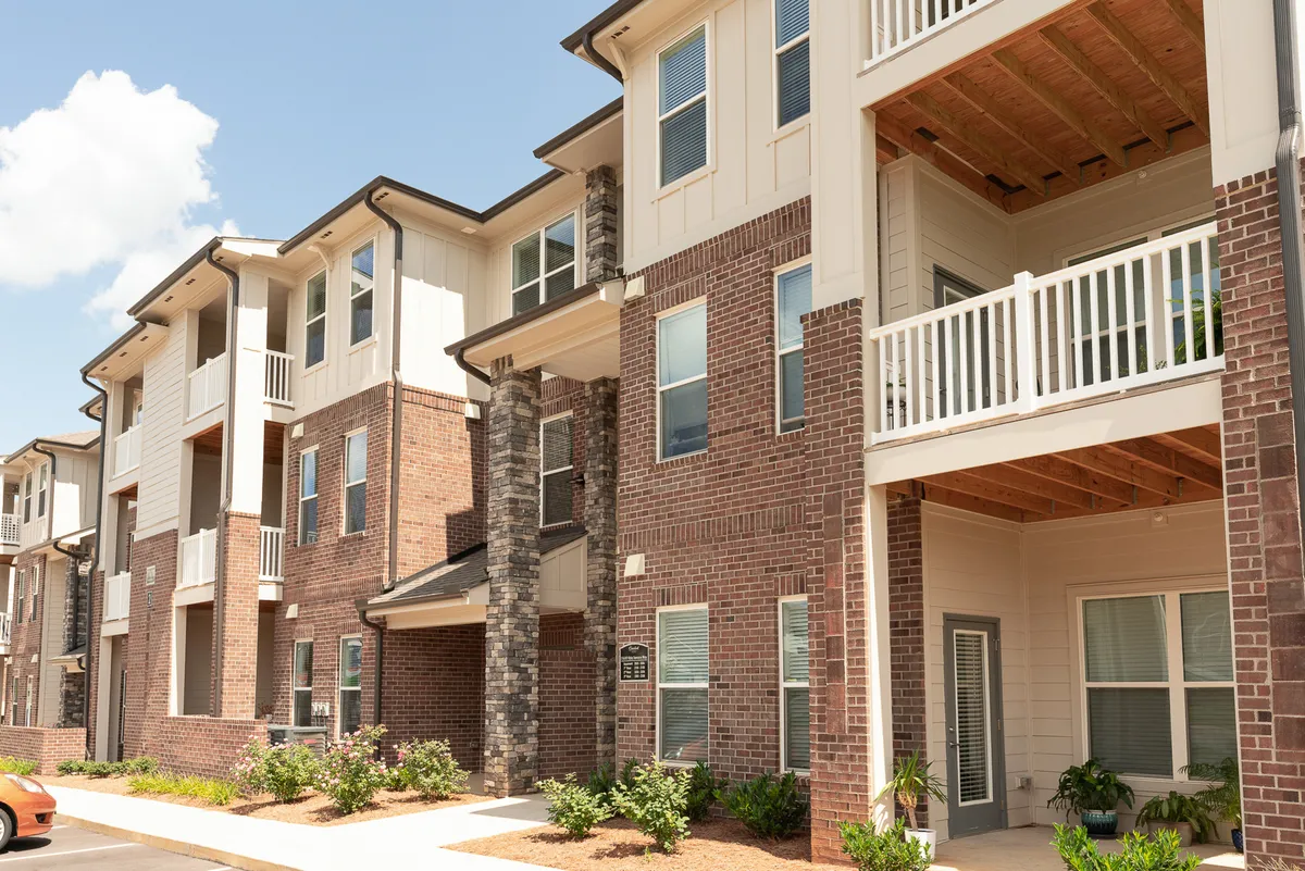 A modern exterior shot featuring the building's brick facade, balconies overlooking landscaped gardens, and inviting communal spaces. Residents are shown enjoying quiet moments on balconies and in the gardens.